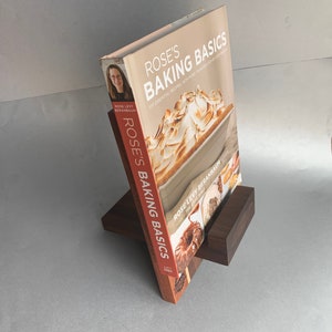 Puede incluir: Un soporte de libro de madera marr&oacute;n con un libro de cocina titulado "Rose's Baking Basics" de Rose Levy Beranbaum. La portada del libro presenta una foto de un postre con una cobertura de merengue.