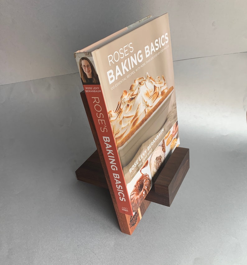 Puede incluir: Un soporte de libro de madera marr&oacute;n con un libro de cocina titulado "Rose's Baking Basics" de Rose Levy Beranbaum. La portada del libro presenta una foto de un postre con una cobertura de merengue.