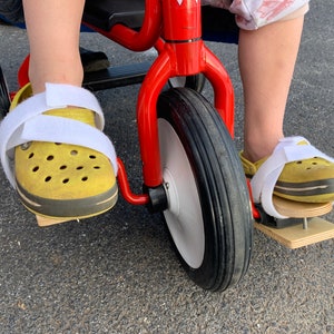 May include: A red tricycle with a white front wheel and black tires. The tricycle has wooden pedals attached to the bottom of the pedals. A person is sitting on the tricycle with their feet on the pedals.