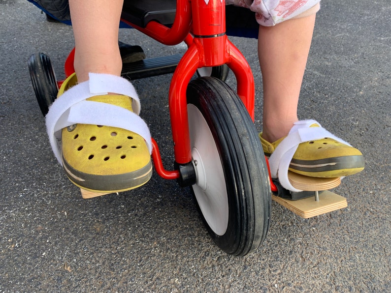 May include: A red tricycle with a white front wheel and black tires. The tricycle has wooden pedals attached to the bottom of the pedals. A person is sitting on the tricycle with their feet on the pedals.
