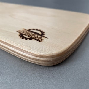 May include: A close-up of the edge of a light wood cutting board with rounded edges. The board is made of plywood and has a laser-etched logo that reads "Jess & Co. Woodworks".
