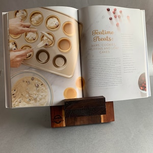 Puede incluir: Libro de cocina abierto con una receta para delicias de la hora del t&eacute;, que incluye barras, galletas, muffins y pasteles de hogaza. El libro est&aacute; apoyado en un soporte de madera. Un taz&oacute;n de masa y un molde para muffins con masa en las tazas son visibles en la p&aacute;gina izquierda.
