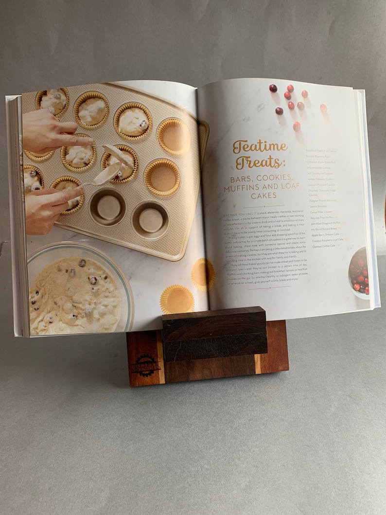 Puede incluir: Libro de cocina abierto con una receta para delicias de la hora del t&eacute;, que incluye barras, galletas, muffins y pasteles de hogaza. El libro est&aacute; apoyado en un soporte de madera. Un taz&oacute;n de masa y un molde para muffins con masa en las tazas son visibles en la p&aacute;gina izquierda.