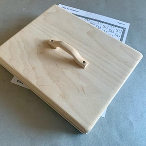 May include: A light-colored wooden box with a handle, possibly a bread box or storage container. The box is rectangular with a rounded handle attached to the top. A pattern is partially visible underneath.