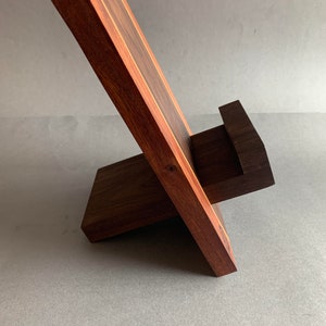 Puede incluir: Un soporte de tel&eacute;fono de madera con una base marr&oacute;n oscuro y una parte superior marr&oacute;n claro. El soporte est&aacute; inclinado para sostener un tel&eacute;fono en posici&oacute;n vertical.