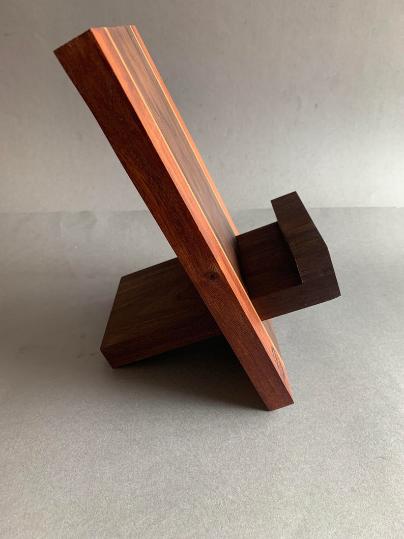 Puede incluir: Un soporte de tel&eacute;fono de madera con una base marr&oacute;n oscuro y una parte superior marr&oacute;n claro. El soporte est&aacute; inclinado para sostener un tel&eacute;fono en posici&oacute;n vertical.
