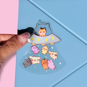 Può includere: Un adesivo trasparente raffigurante un'astronave dei cartoni animati con un orso pilota e diversi animali. L'adesivo è tenuto sopra un quaderno azzurro. Il testo "Rakuten Kobo" è visibile.