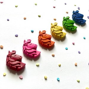 May include: A set of seven unicorn-shaped crayons in a rainbow of colors, arranged in a line on a white background with colorful confetti.
