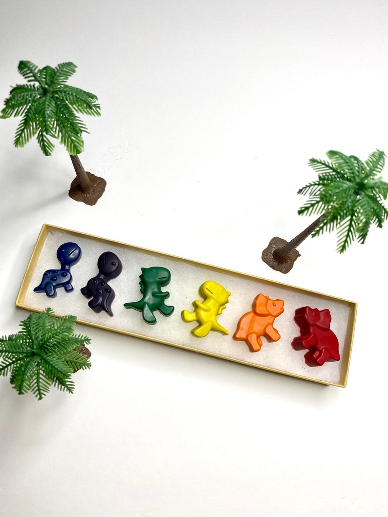 May include: A set of six dinosaur-shaped crayons in a rainbow of colors, including red, orange, yellow, green, blue, and purple. The crayons are arranged in a row in a box with a white background.