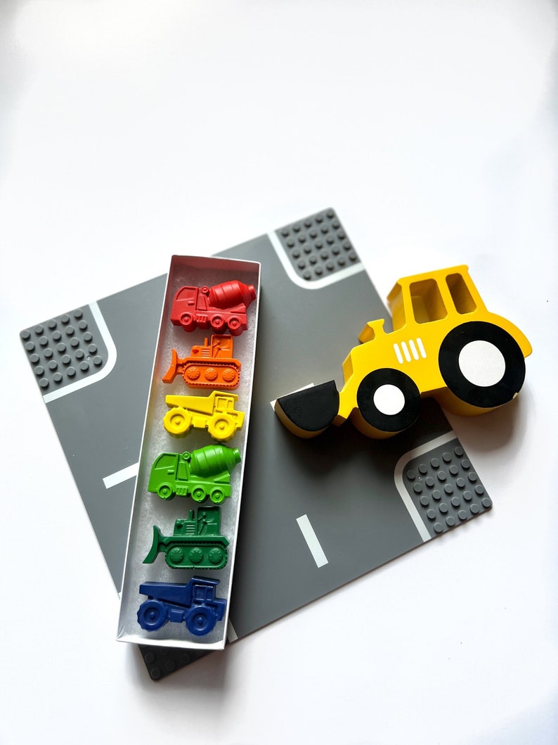 May include: A set of construction vehicle crayons in a rainbow of colors, including a yellow bulldozer, a red cement mixer, and a green dump truck. The crayons are in a clear plastic box with a white background. A yellow toy construction vehicle is on a gray play mat with black Lego-like blocks.