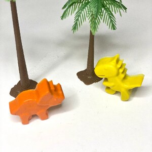 May include: Two toy dinosaurs, one orange and one yellow, are placed on a white surface in front of two small palm trees.