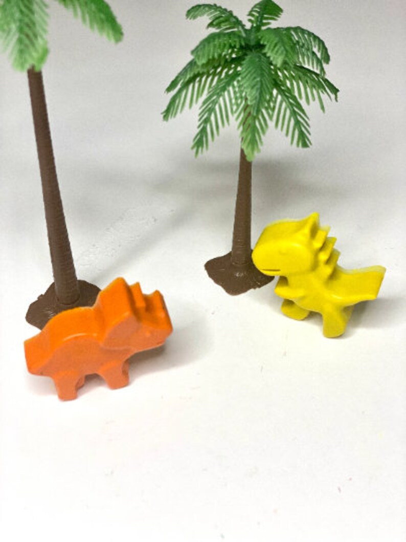 May include: Two toy dinosaurs, one orange and one yellow, are placed on a white surface in front of two small palm trees.