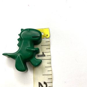 May include: A green plastic dinosaur toy measuring approximately 1 inch tall. The dinosaur is standing on a white surface next to a measuring tape.
