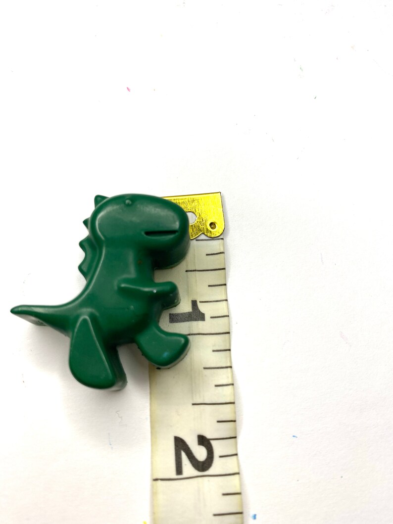 May include: A green plastic dinosaur toy measuring approximately 1 inch tall. The dinosaur is standing on a white surface next to a measuring tape.