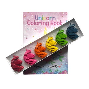 May include: A rectangular box containing six unicorn-shaped crayons in red, pink, orange, yellow, green, and blue, positioned diagonally over a Unicorn Coloring Book with a pastel background featuring stars, moons, and floral unicorn illustrations.
