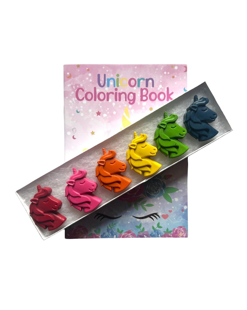 May include: A rectangular box containing six unicorn-shaped crayons in red, pink, orange, yellow, green, and blue, positioned diagonally over a Unicorn Coloring Book with a pastel background featuring stars, moons, and floral unicorn illustrations.