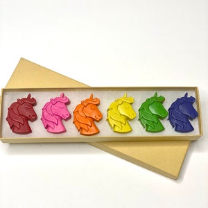 May include: A set of six unicorn-shaped crayons in a rainbow of colors: red, pink, orange, yellow, green, and blue. The crayons are arranged in a box with a brown lid.
