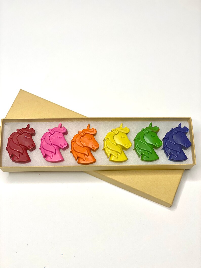 May include: A set of six unicorn-shaped crayons in a rainbow of colors: red, pink, orange, yellow, green, and blue. The crayons are arranged in a box with a brown lid.