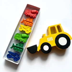 May include: A set of six construction vehicle crayons in a clear box. The crayons are arranged in a rainbow order from red to blue. The crayons are shaped like a cement mixer, bulldozer, dump truck, backhoe, and two different types of trucks. A yellow wooden toy construction vehicle is also pictured.