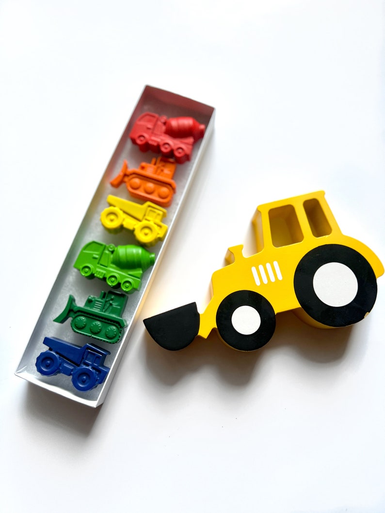 May include: A set of six construction vehicle crayons in a clear box. The crayons are arranged in a rainbow order from red to blue. The crayons are shaped like a cement mixer, bulldozer, dump truck, backhoe, and two different types of trucks. A yellow wooden toy construction vehicle is also pictured.