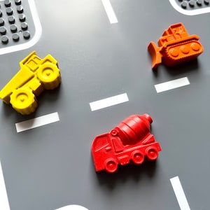 May include: A gray play mat with white lines resembling a road. Three toy construction vehicles are placed on the mat: a yellow dump truck, a red cement mixer, and an orange bulldozer.