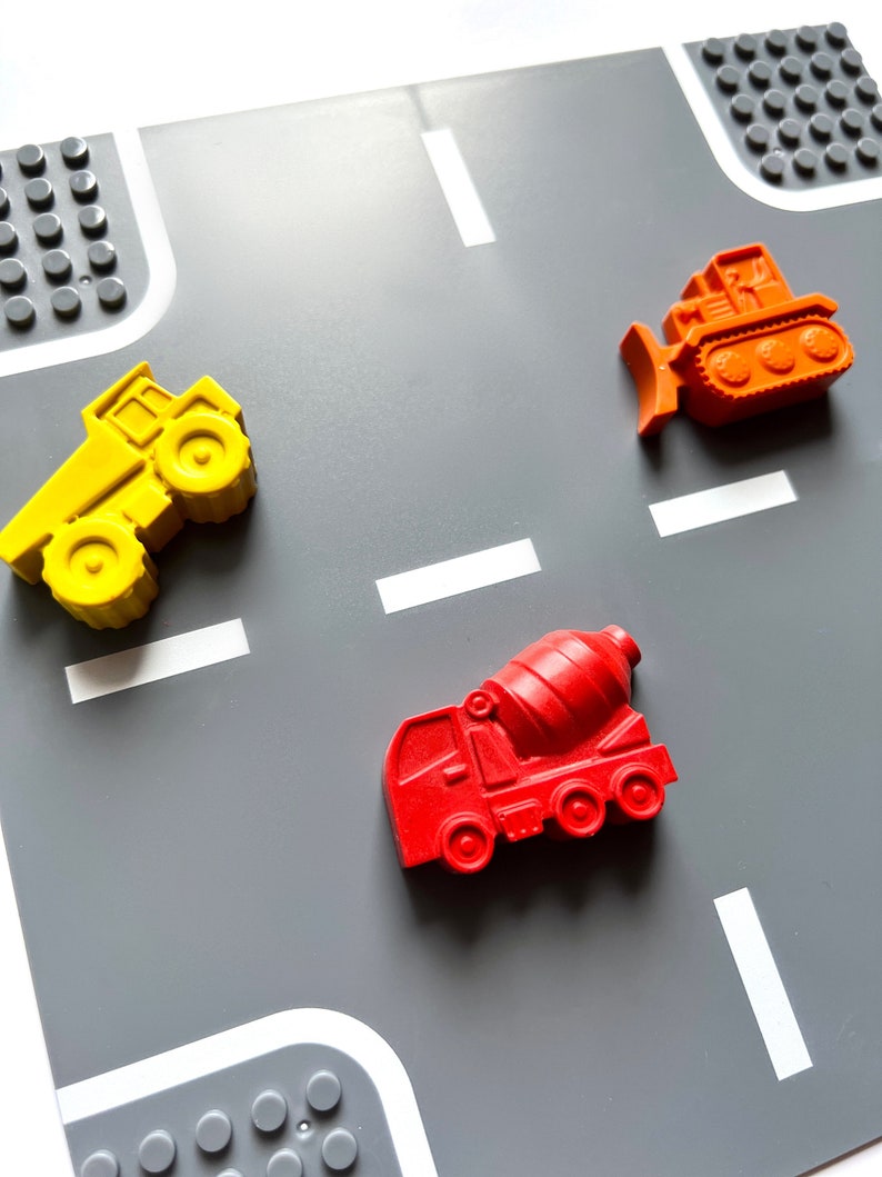 May include: A gray play mat with white lines resembling a road. Three toy construction vehicles are placed on the mat: a yellow dump truck, a red cement mixer, and an orange bulldozer.