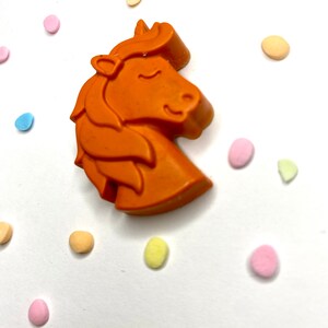 May include: Orange unicorn shaped candle with a white background and colorful confetti.