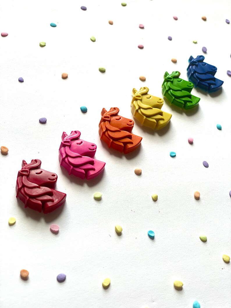May include: A set of seven unicorn-shaped crayons in a rainbow of colors, arranged in a line on a white background with colorful confetti.