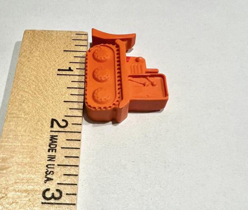 May include: Orange plastic toy bulldozer measuring approximately 2 inches in length. The bulldozer has a detailed design with tracks and a raised blade.