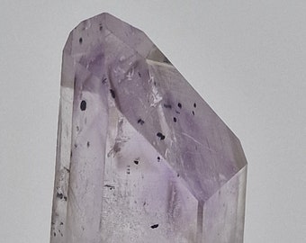 May include: A clear, light purple amethyst crystal with black inclusions. The crystal is pointed at the top and has a rough, gray base.