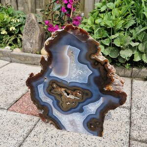 May include: A large, natural agate slice with brown, white and blue banding. The agate is polished and has a smooth surface. It is sitting on a grey concrete surface.