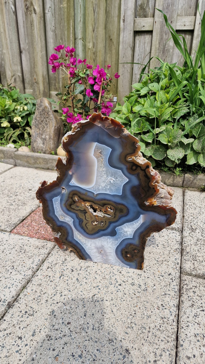 May include: A large, natural agate slice with brown, white and blue banding. The agate is polished and has a smooth surface. It is sitting on a grey concrete surface.