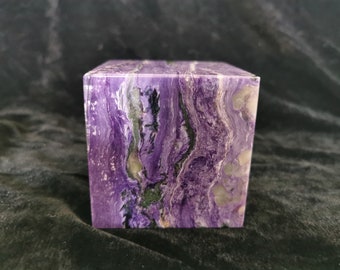 May include: A square stone block with a polished surface, displaying a swirling pattern of purple, white, and green. The cube-shaped object is likely a decorative item or a piece of art. The stone's measurements are approximately 7 cm x 7 cm x 7 cm.