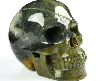 Puede incluir: Una escultura de calavera de ágata verde y marrón. La calavera está tallada con detalles precisos, incluidos los dientes y las cuencas de los ojos.