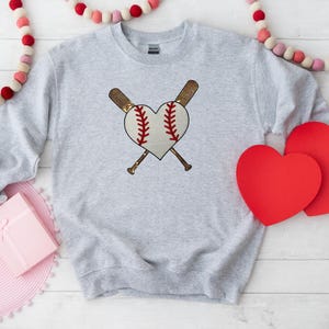 May include: A gray sweatshirt with a baseball and crossed bats design in the shape of a heart. The design is in red, white, and gold.
