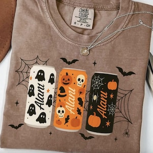 May include: A brown Comfort Colors t-shirt with a Halloween-themed design. The shirt features three stylized beverage cans with the word "Alani" on each. The cans are decorated with ghosts, pumpkins, spiders, and bats. A silver necklace is draped across the shirt.