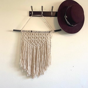 May include: A white macrame wall hanging with fringe, hanging from a wooden dowel rod on a white wall. A burgundy fedora hat is hanging on a hook above the wall hanging.