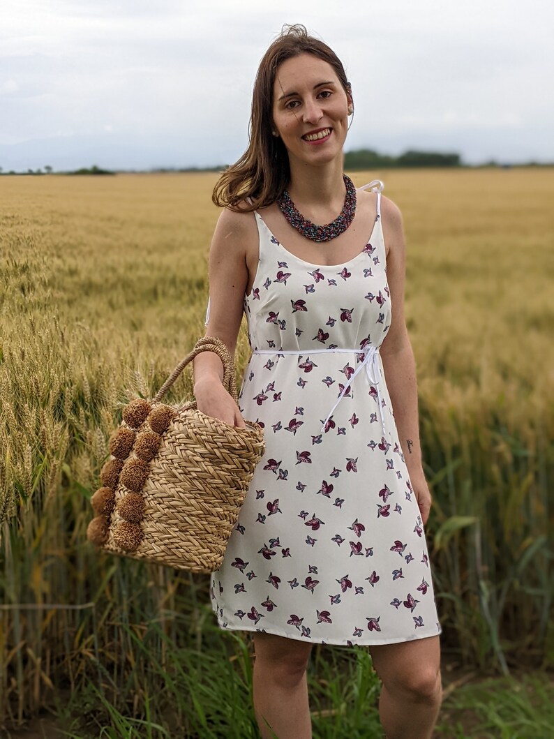 May include: A woman wearing a white dress with a floral pattern and a beaded necklace, standing in a field of wheat, holding a straw bag with pom-poms.