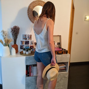 May include: A woman wearing a white satin tank top, denim shorts, and white ankle boots. She is holding a straw hat and standing in front of a white dresser with a record player on top.