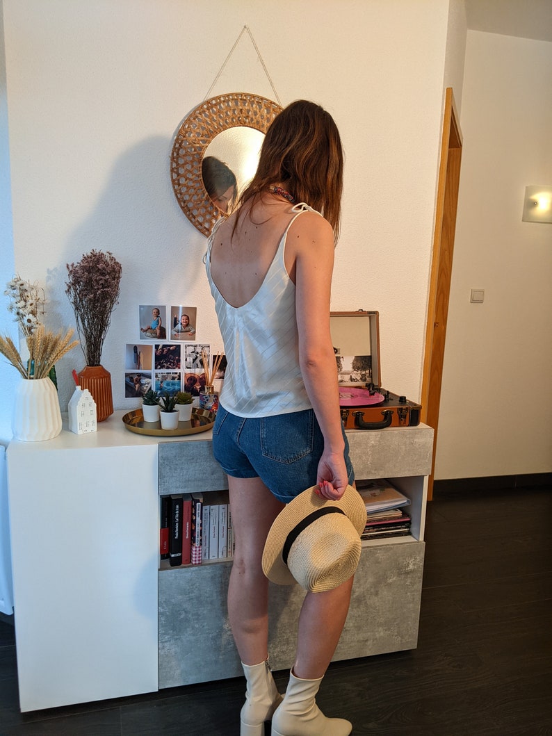 May include: A woman wearing a white satin tank top, denim shorts, and white ankle boots. She is holding a straw hat and standing in front of a white dresser with a record player on top.