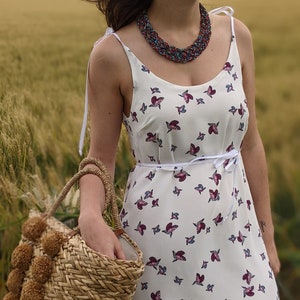 May include: A woman wearing a white sundress with a floral print and a multi-colored beaded necklace. She is holding a straw tote bag with brown pom-poms.