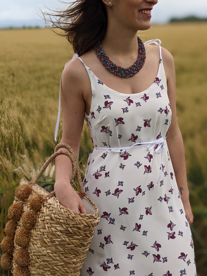 May include: A woman wearing a white sundress with a floral print and a multi-colored beaded necklace. She is holding a straw tote bag with brown pom-poms.