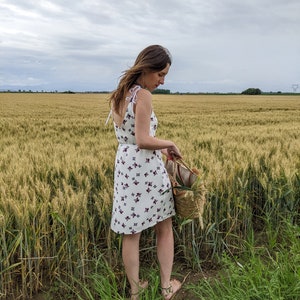 May include: A woman in a white dress with a floral pattern stands in a field of wheat. She is holding a wicker basket.