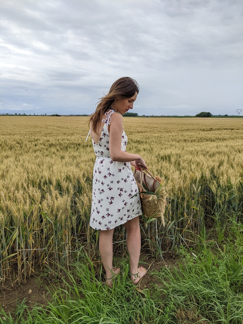 May include: A woman in a white dress with a floral pattern stands in a field of wheat. She is holding a wicker basket.