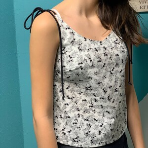 May include: A white tank top with a black floral print and adjustable straps. The straps are tied at the shoulders.