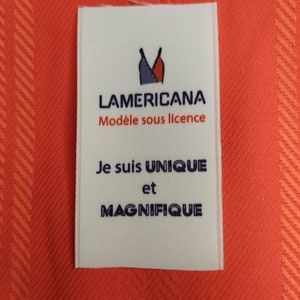 Peut inclure: Étiquette de vêtement blanche avec un logo bleu et rouge d'un haut de maillot de bain pour femme. Le texte "LAMERICANA" est en bleu, suivi de "Modèle sous licence" en noir. En dessous, le texte "Je suis UNIQUE et MAGNIFIQUE" en bleu.