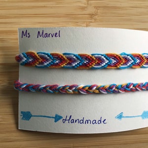 May include: Two handmade friendship bracelets with a chevron pattern in red, orange, yellow, blue, white, and purple. The bracelets are displayed on a white card with the text "Ms Marvel" and "Handmade" written in blue ink.