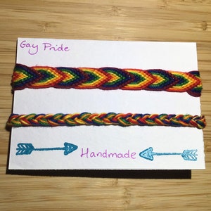 May include: Two handmade friendship bracelets in rainbow colours with the text "Gay Pride" and "Handmade" on a white card.