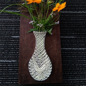 May include: A white string art vase with a brown wooden background. The vase is filled with orange flowers and green leaves.