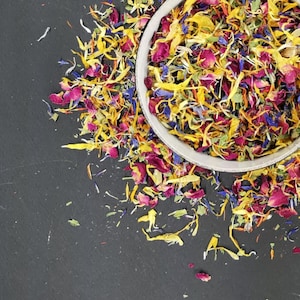 Può includere: Una ciotola bianca piena di un mix di petali di fiori secchi in tonalità di giallo, rosa, viola e verde. I petali sono sparsi intorno alla ciotola su una superficie nera.